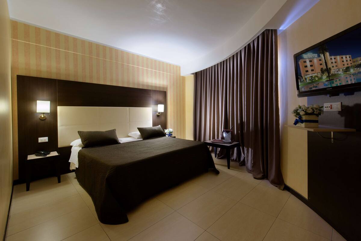Habitación de hotel con una cama grande, una mesita de noche, un escritorio con lámpara, un televisor en la pared y una cortina.
