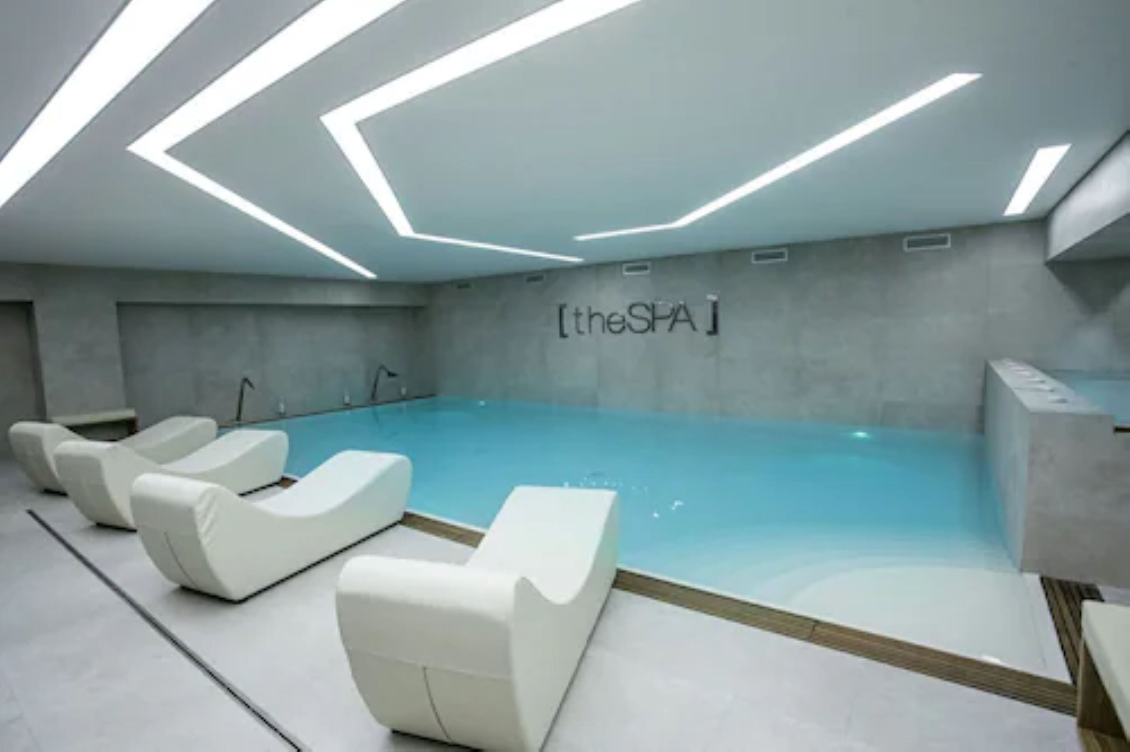 A modern indoor swimming pool with a geometric ceiling design and lounge chairs.