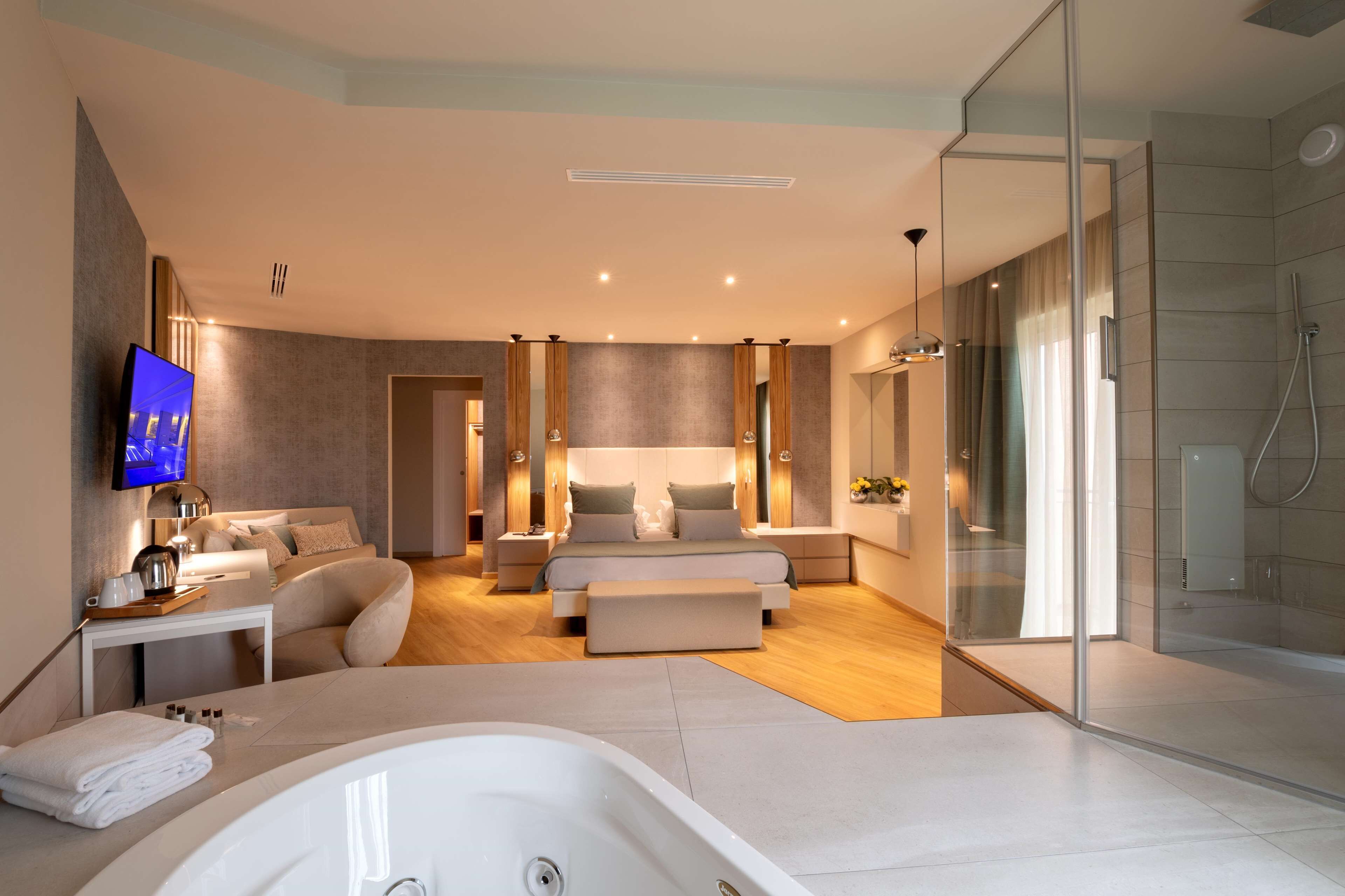 Quarto moderno com cama grande, mesas de cabeceira, televisão e área de chuveiro com vidro.