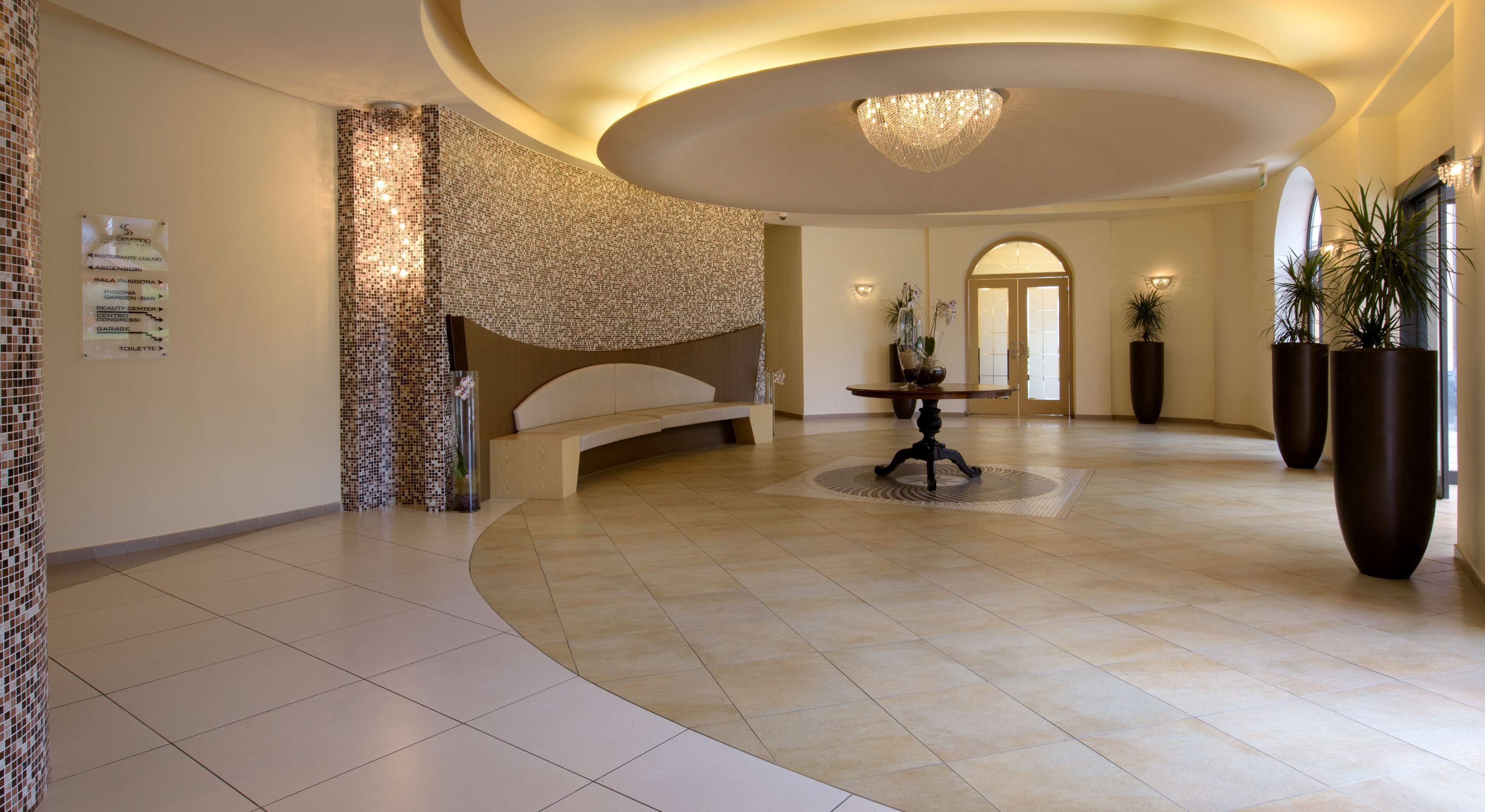 A spacious lobby with a curved wall, a central table, and large potted plants.
