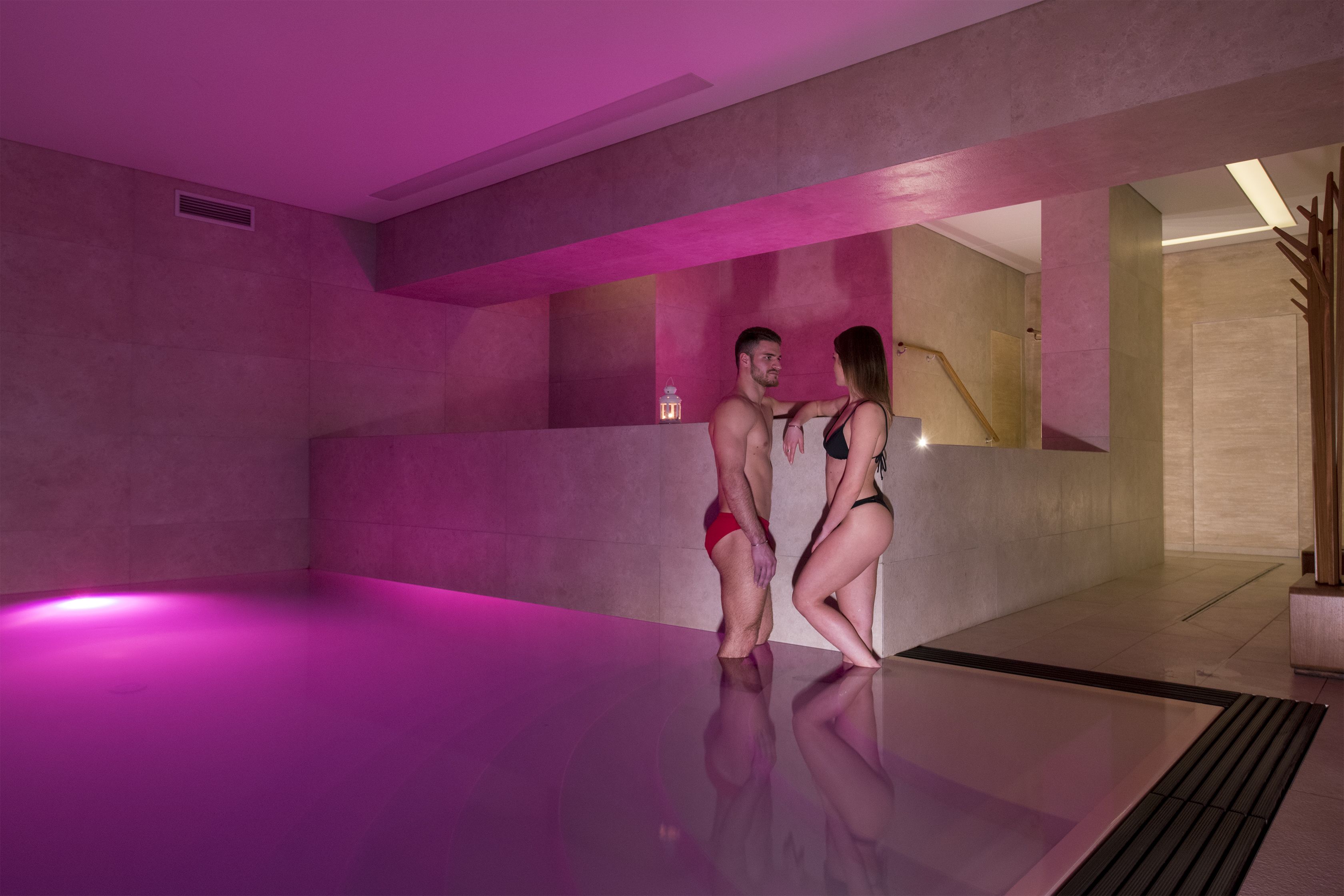 Una piscina interior moderna con diseño minimalista, que destaca por su gran superficie reflectante y una iluminación ambiental en tonos rosados.