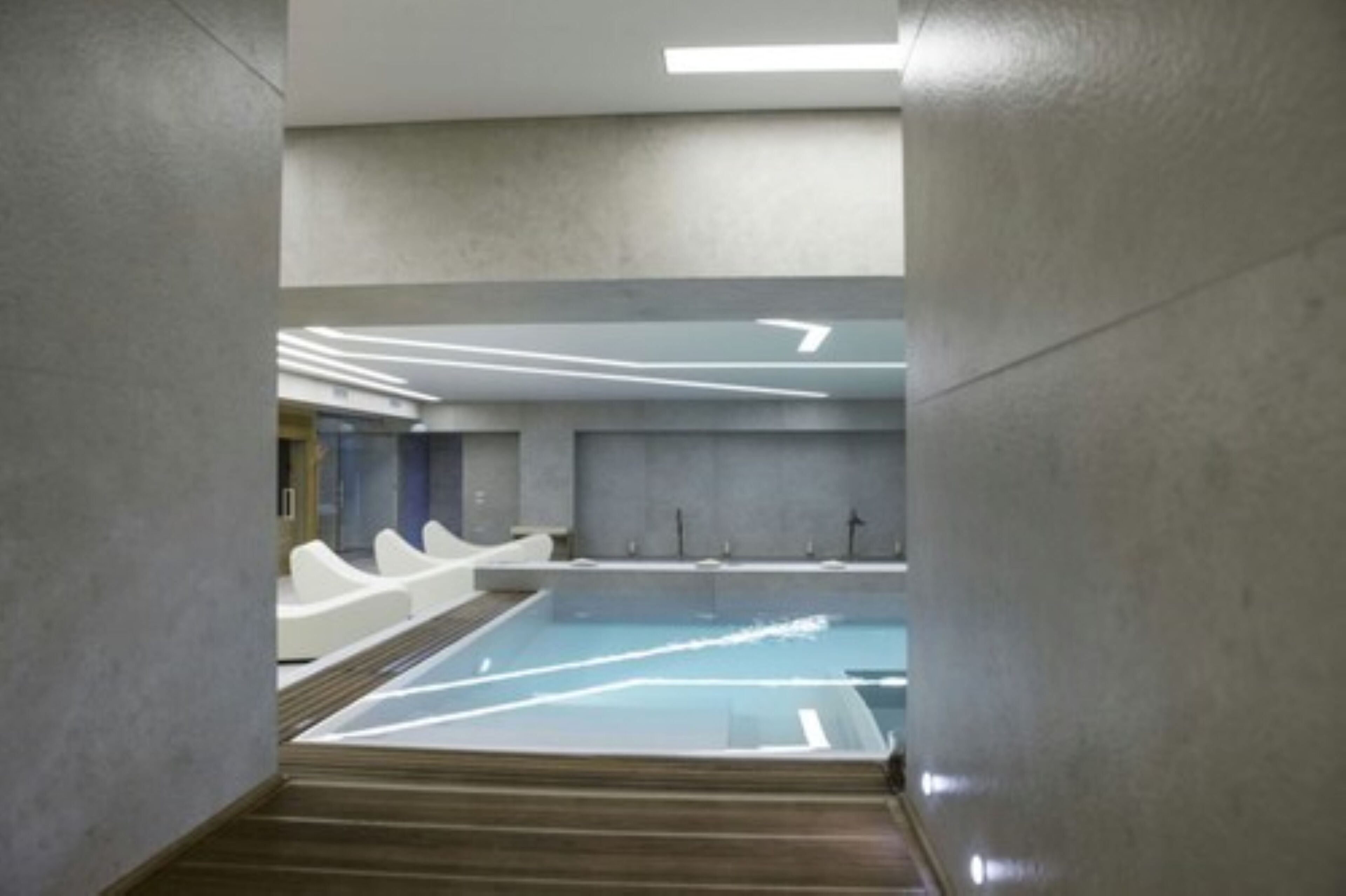 A modern indoor swimming pool with a clear water surface, surrounded by grey walls and wooden flooring.