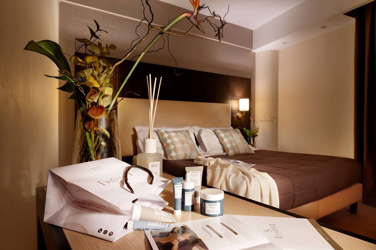Una habitación de hotel con una cama bien hecha, una mesita de noche con artículos de aseo, un florero con flores y una lámpara.