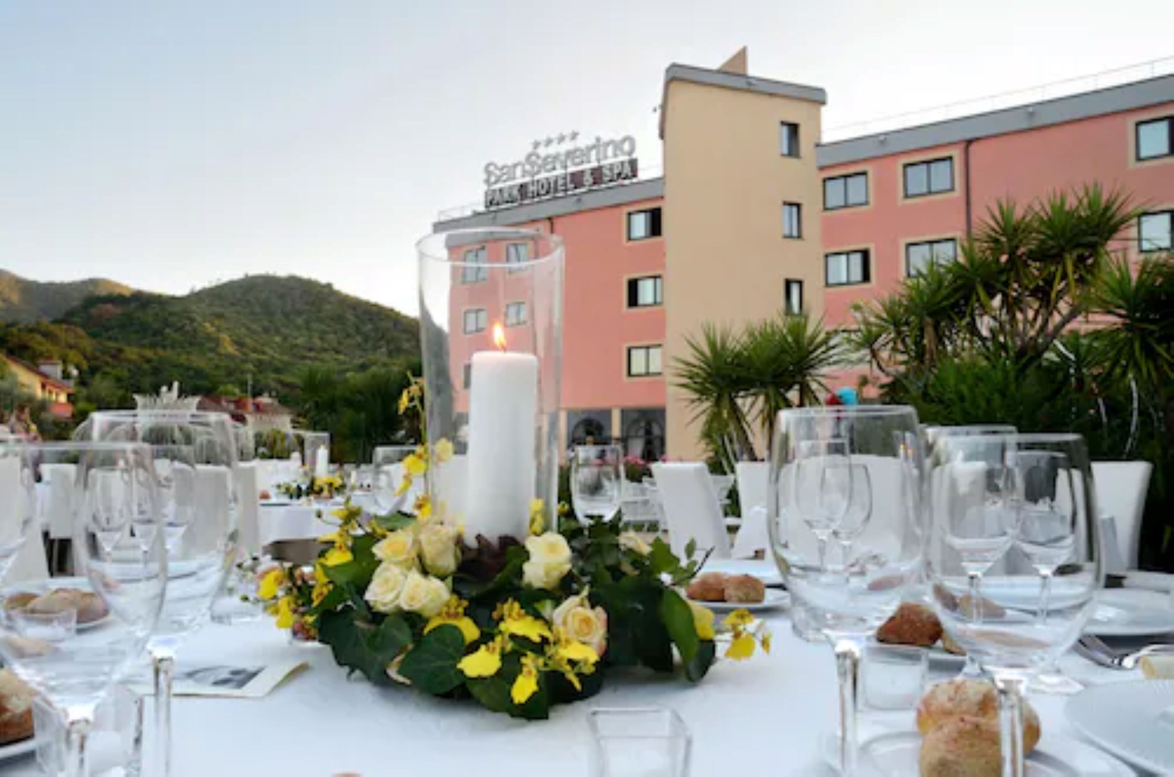 Una mesa dispuesta para una comida, con un centro de mesa con velas, copas transparentes y una vista de un edificio de hotel al fondo.