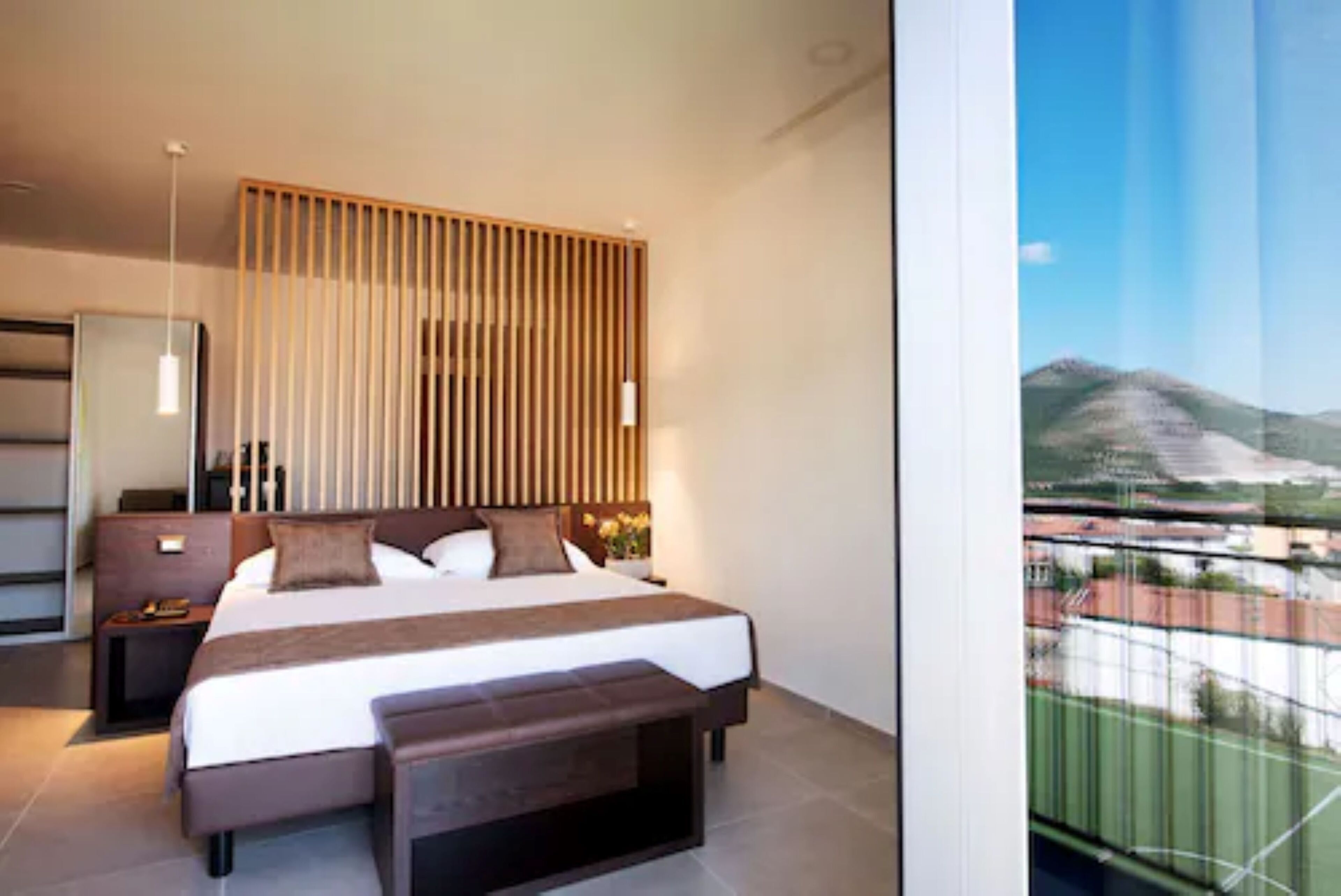 Une chambre d’hôtel moderne avec un grand lit, une table de chevet, une commode et une vue sur un paysage montagneux par une grande fenêtre.