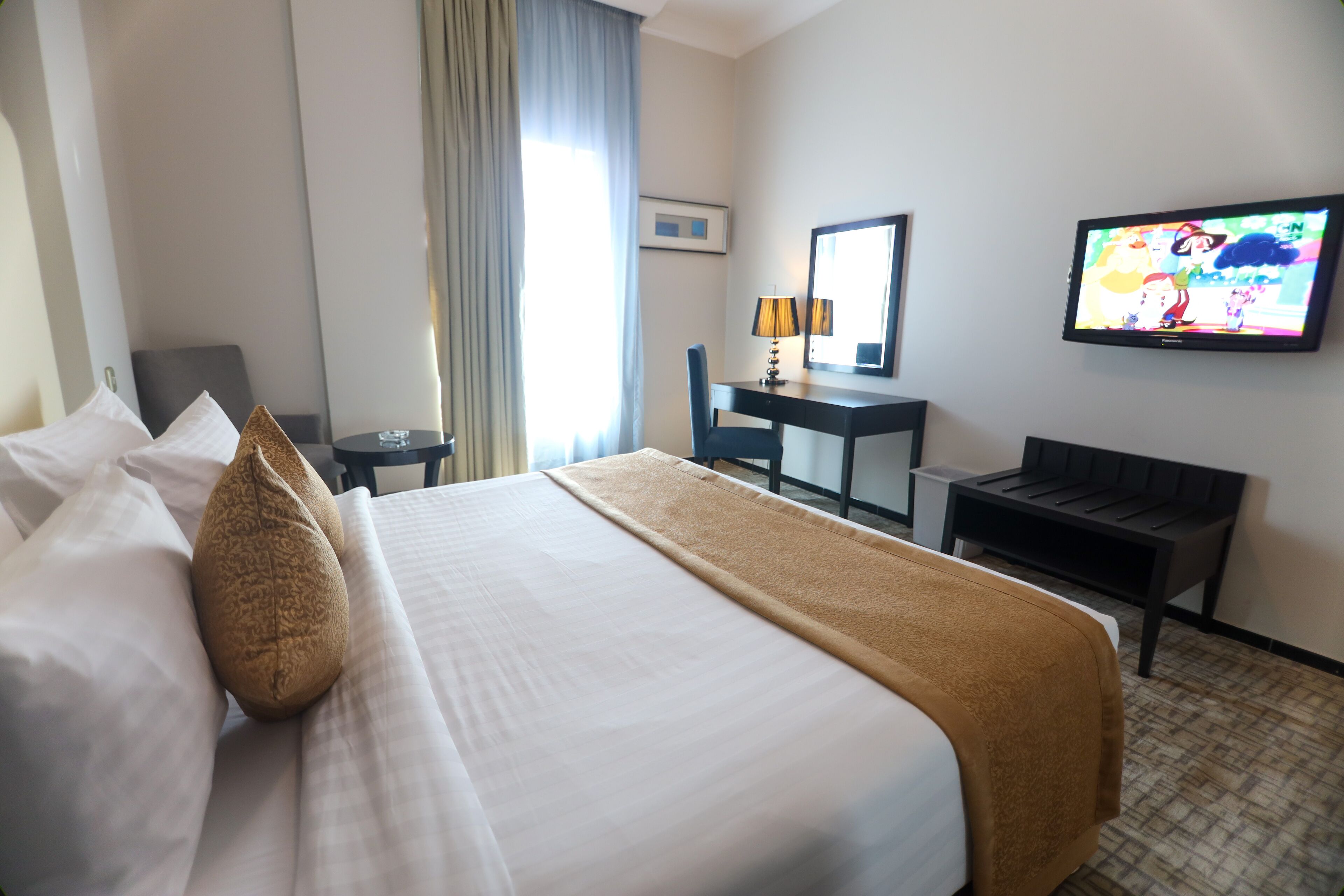 A hotel room with a bed, a desk, a chair, a TV, and a window with curtains.
