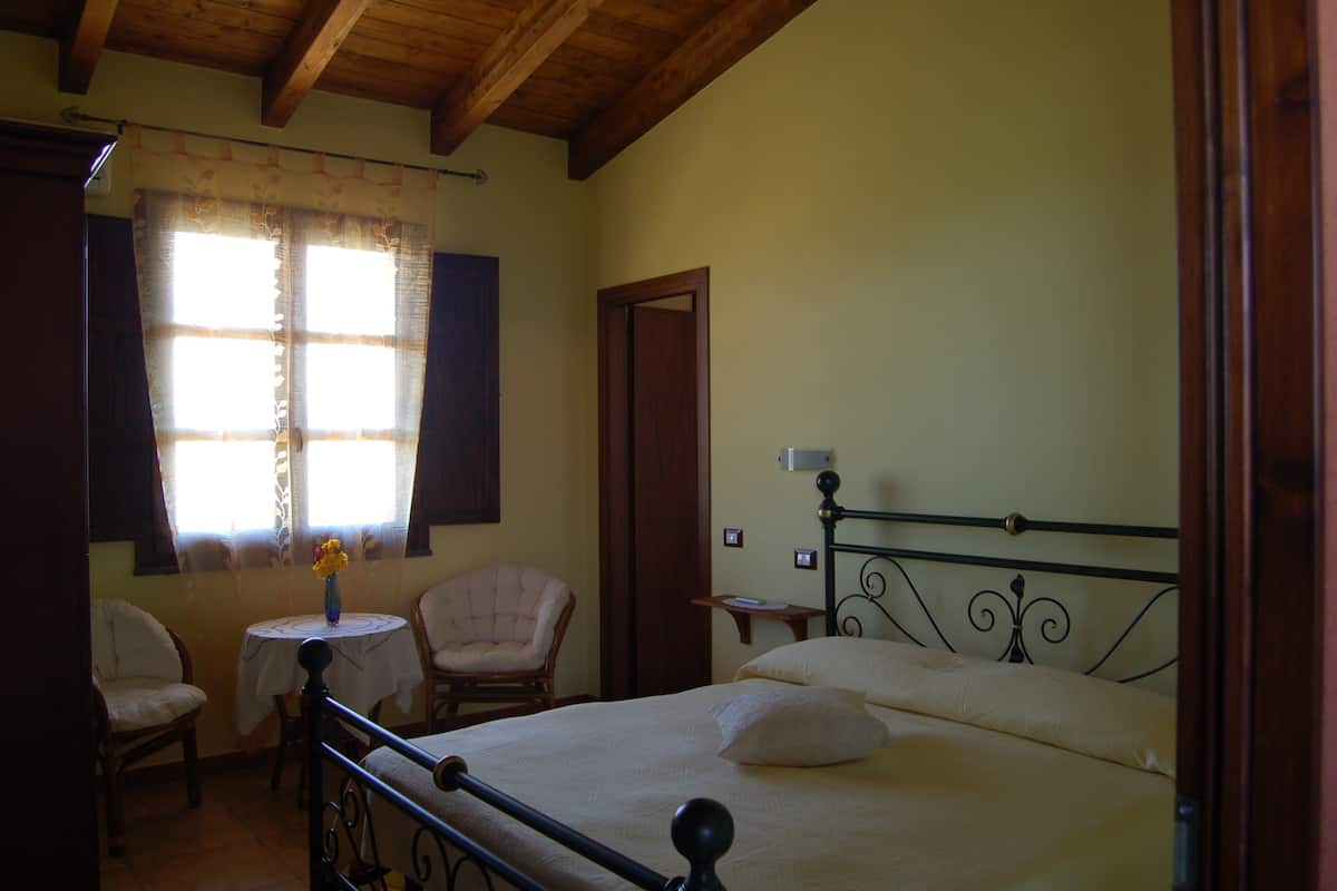 Un dormitorio con una cama, dos sillas, una mesita con un florero y una ventana con cortinas.