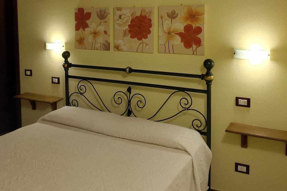 Una cama con cubrecama blanco, cabecera de metal y tres cuadros de flores en la pared.