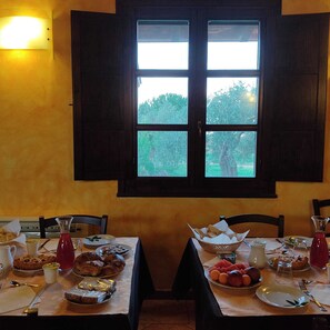 Una mesa de comedor con comida y bebidas, una ventana con contraventanas y una lámpara fijada a la pared.