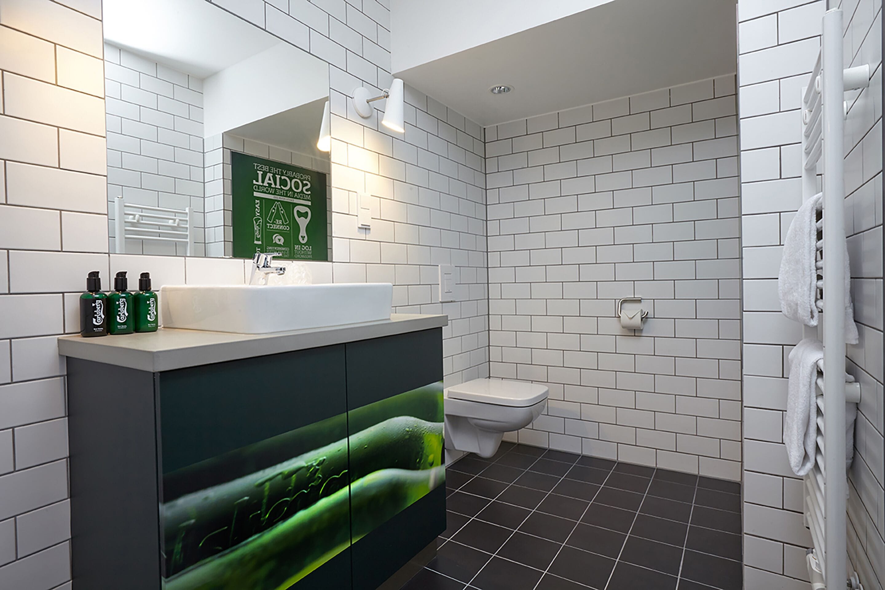 A modern bathroom with white tiles, a dark floor, a white toilet, a sink with green bottles, and a towel rack.