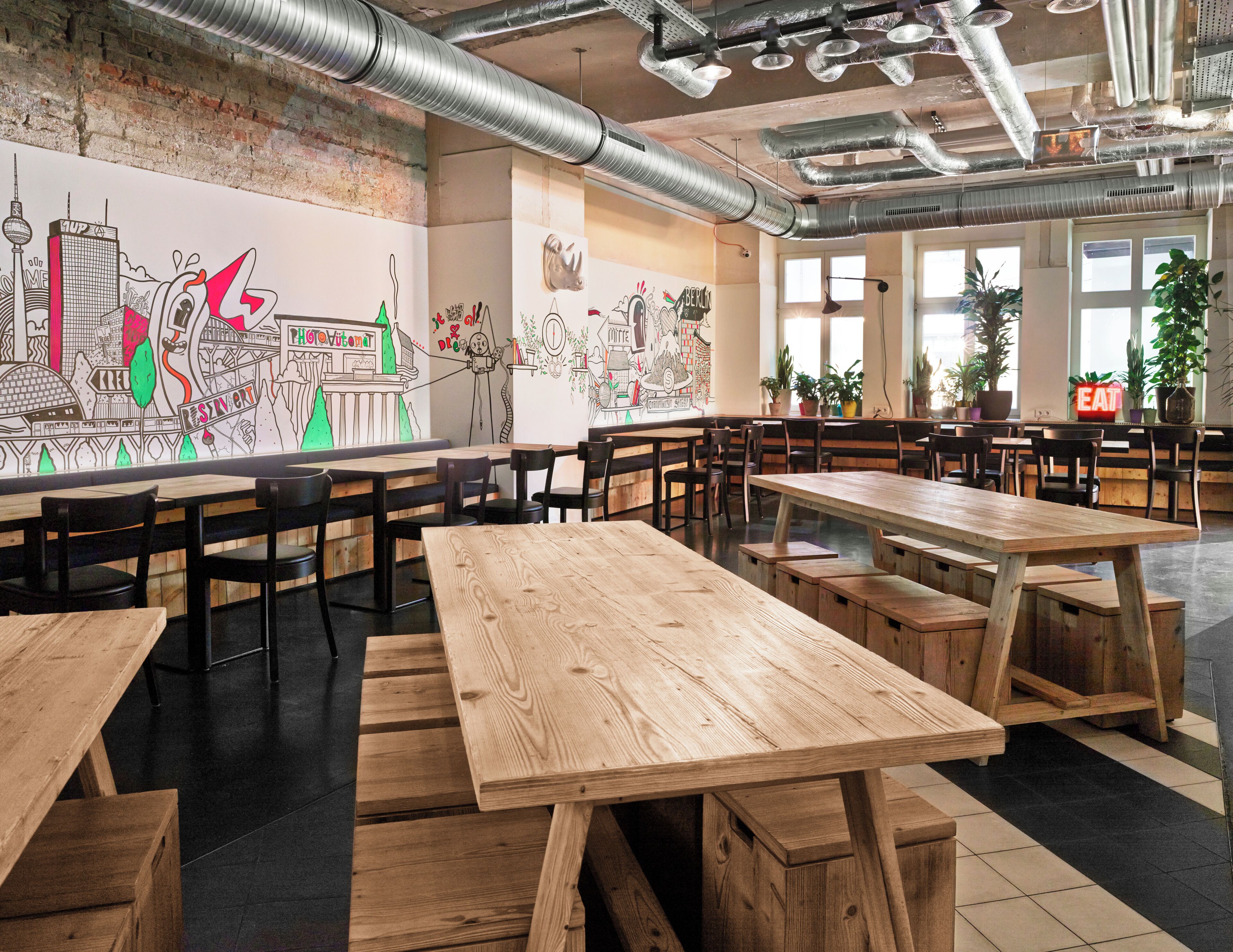 A spacious dining area with wooden tables and chairs, a mural on the wall, and large windows allowing natural light.