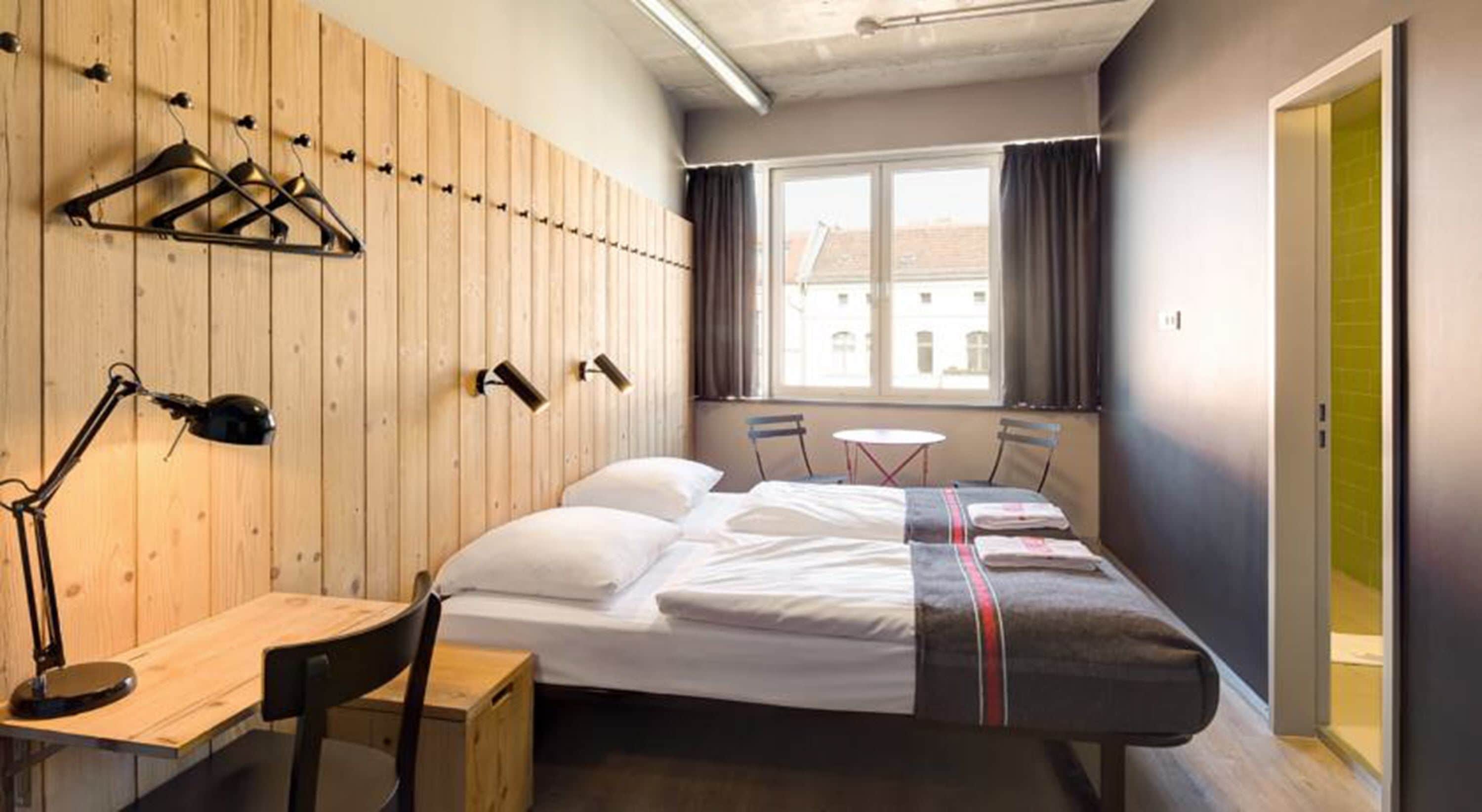 Habitación de hotel con pared de madera, una cama con manta a rayas, un escritorio con lámpara, una mesita y una ventana con cortinas.