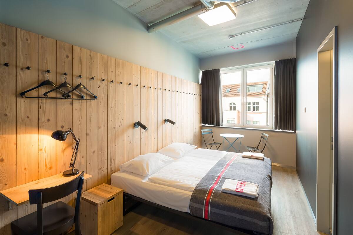 A hotel room with a wooden wall, a bed with a grey and red striped blanket, a desk with a chair, a small table, and a window with curtains.