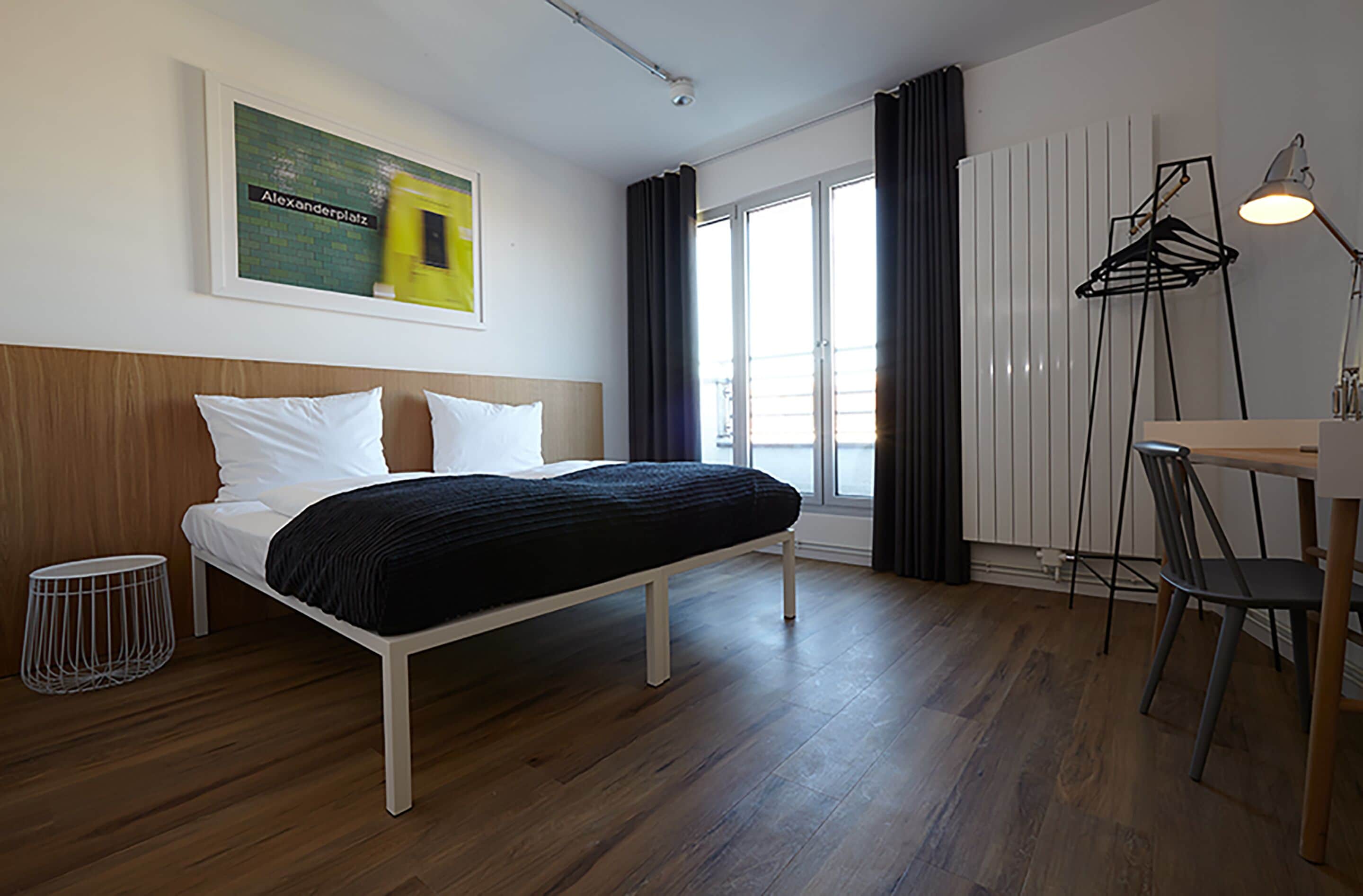 Una habitación de hotel moderna con una cama de madera, una pequeña mesa con una silla, una lámpara de pie y un gran ventanal con cortinas.