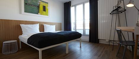 Una habitación de hotel moderna con una cama de madera, una pequeña mesa con una silla, una lámpara de pie y un gran ventanal con cortinas.