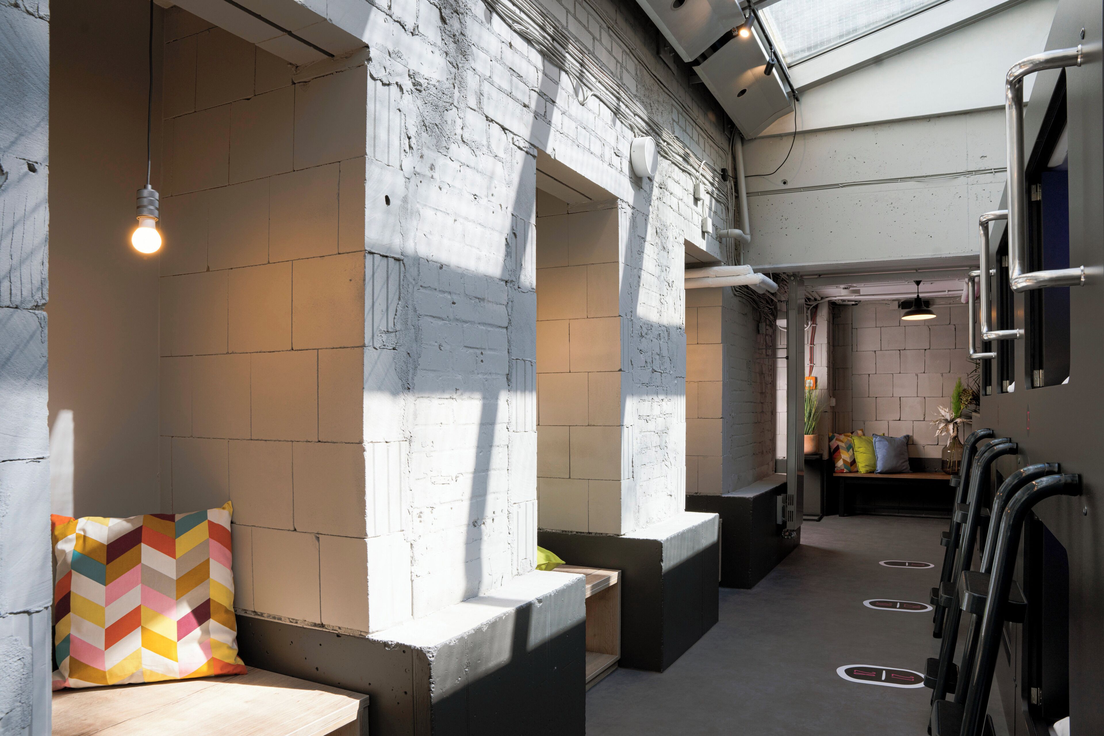 A modern interior with white brick walls, a seating area with a colorful cushion, and a row of black luggage racks.