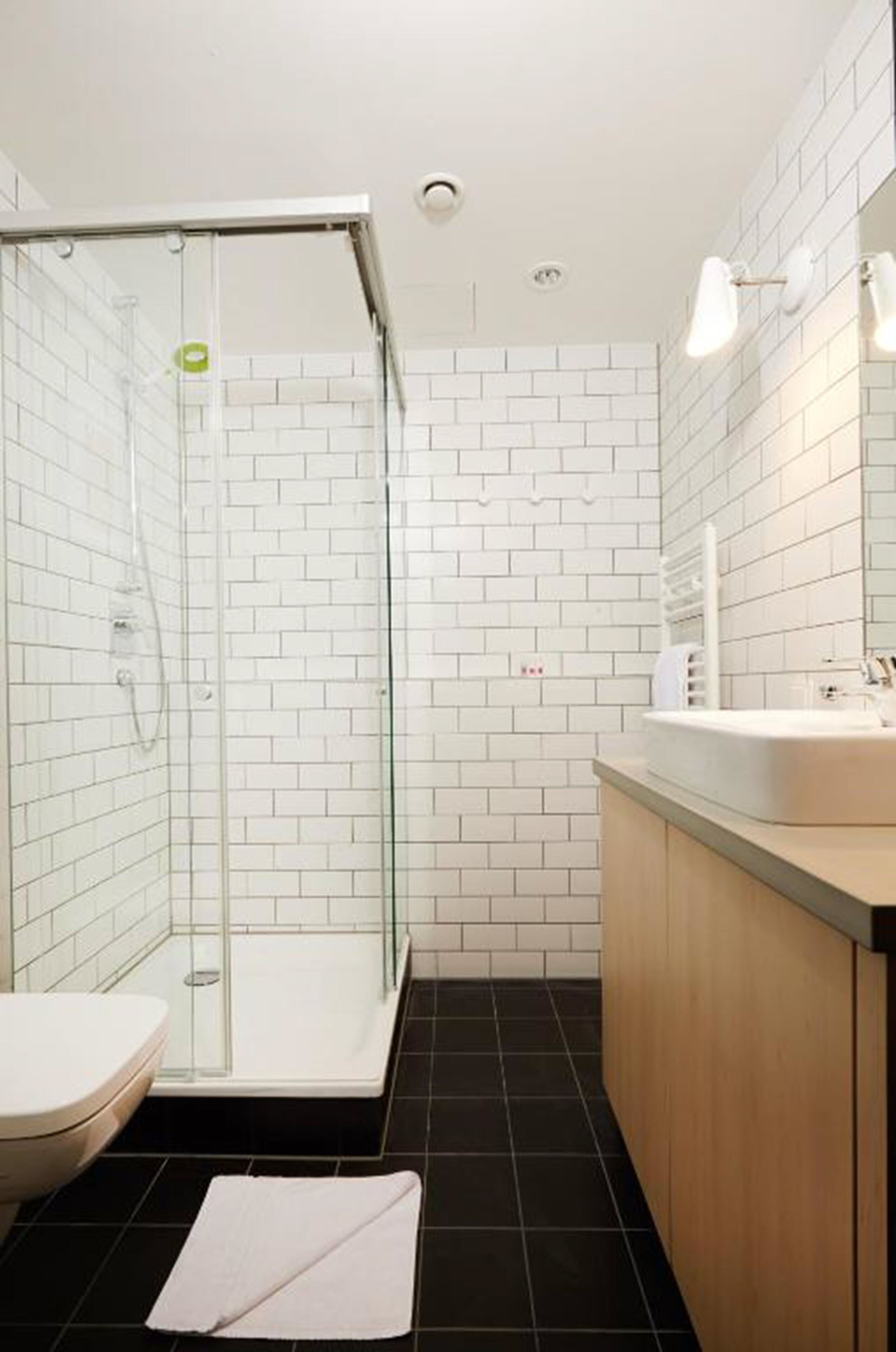 Un baño moderno con azulejos blancos tipo metro, ducha con cerramiento de vidrio, lavamanos blanco y mueble de tocador de madera.