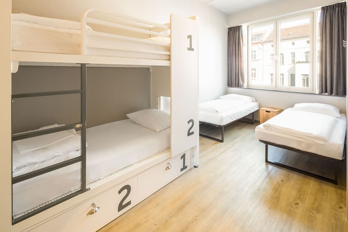 A dormitory room with bunk beds, a single bed, and a window with curtains.