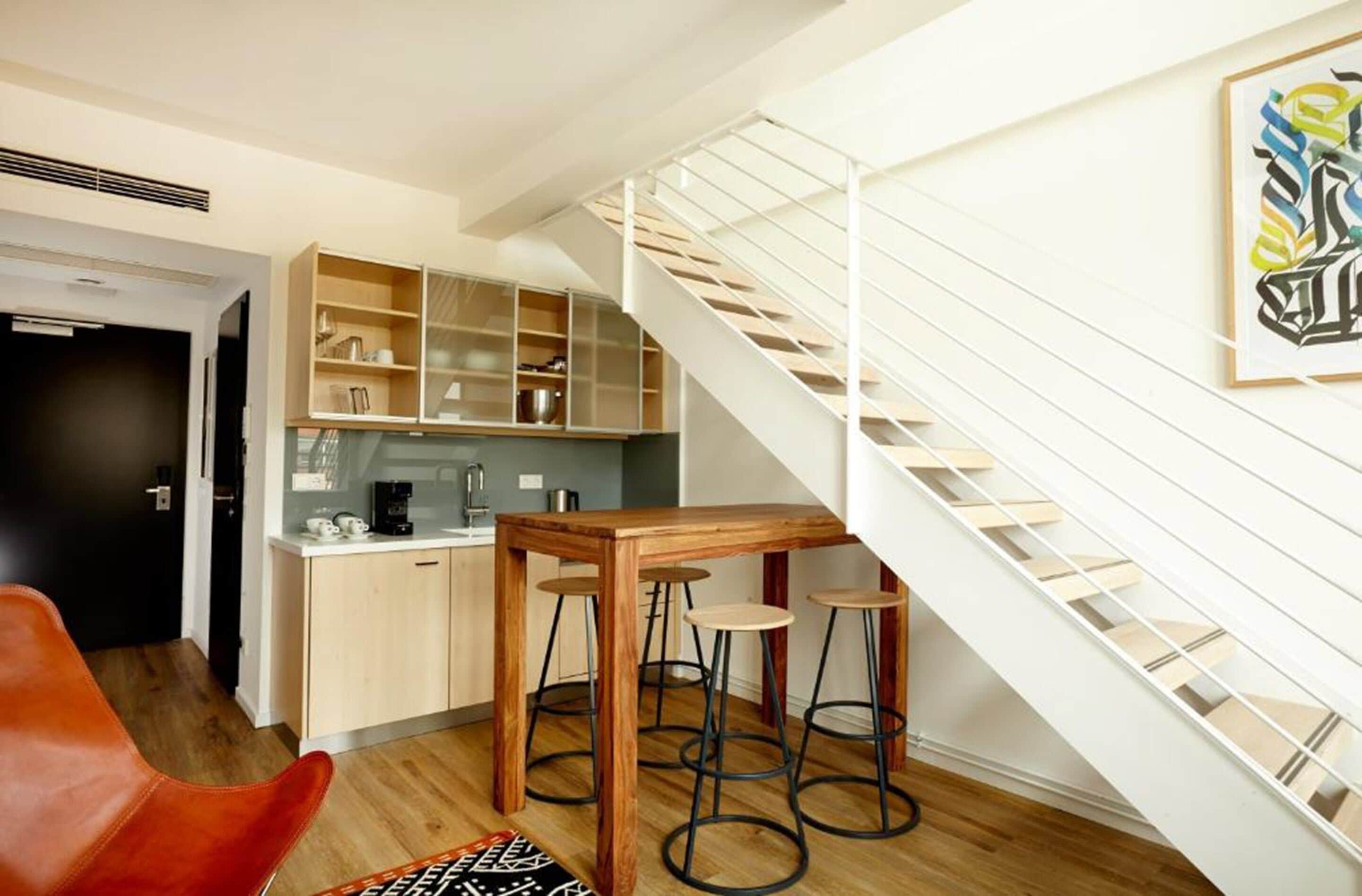 A modern kitchen with a wooden table and stools, a staircase, and a framed artwork on the wall.