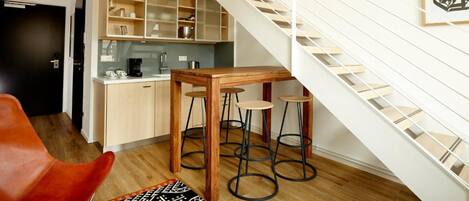 A modern kitchen with a wooden table and stools, a staircase, and a framed artwork on the wall.