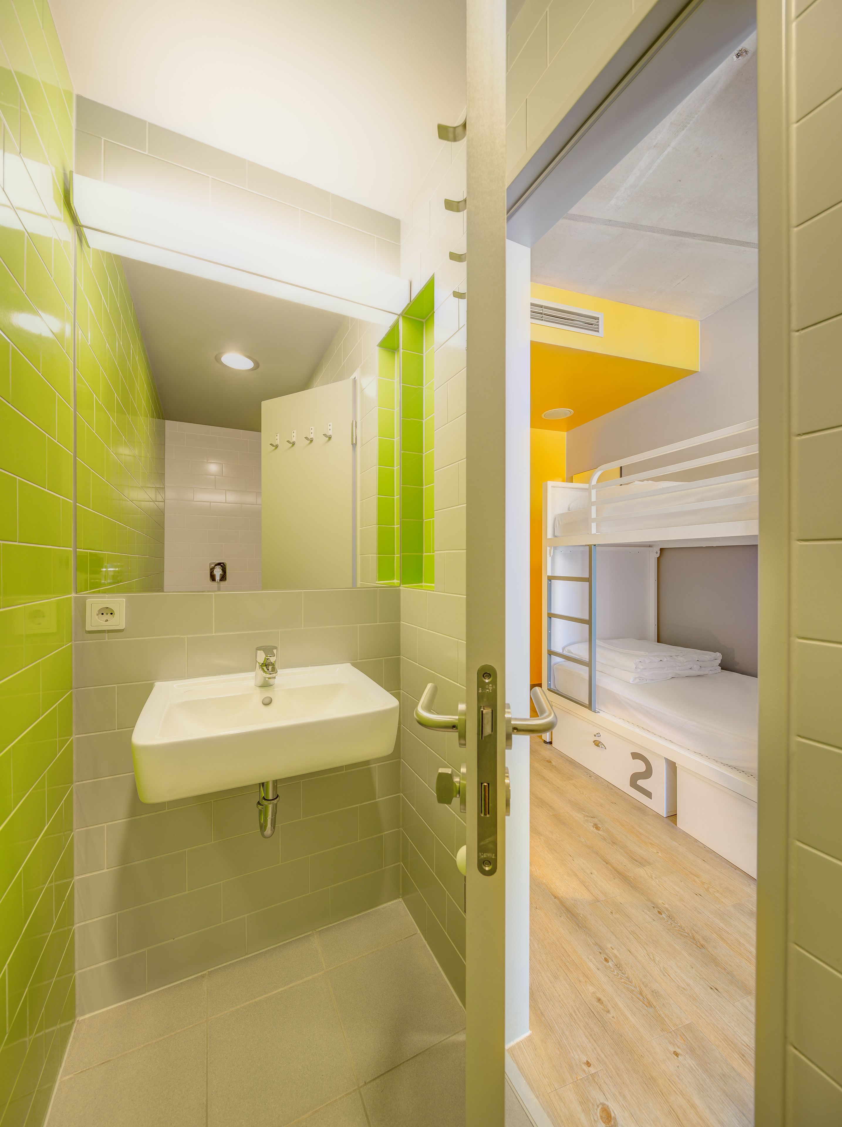A bathroom with a sink, mirror, and green-tiled walls. There is a door leading to a room with bunk beds.