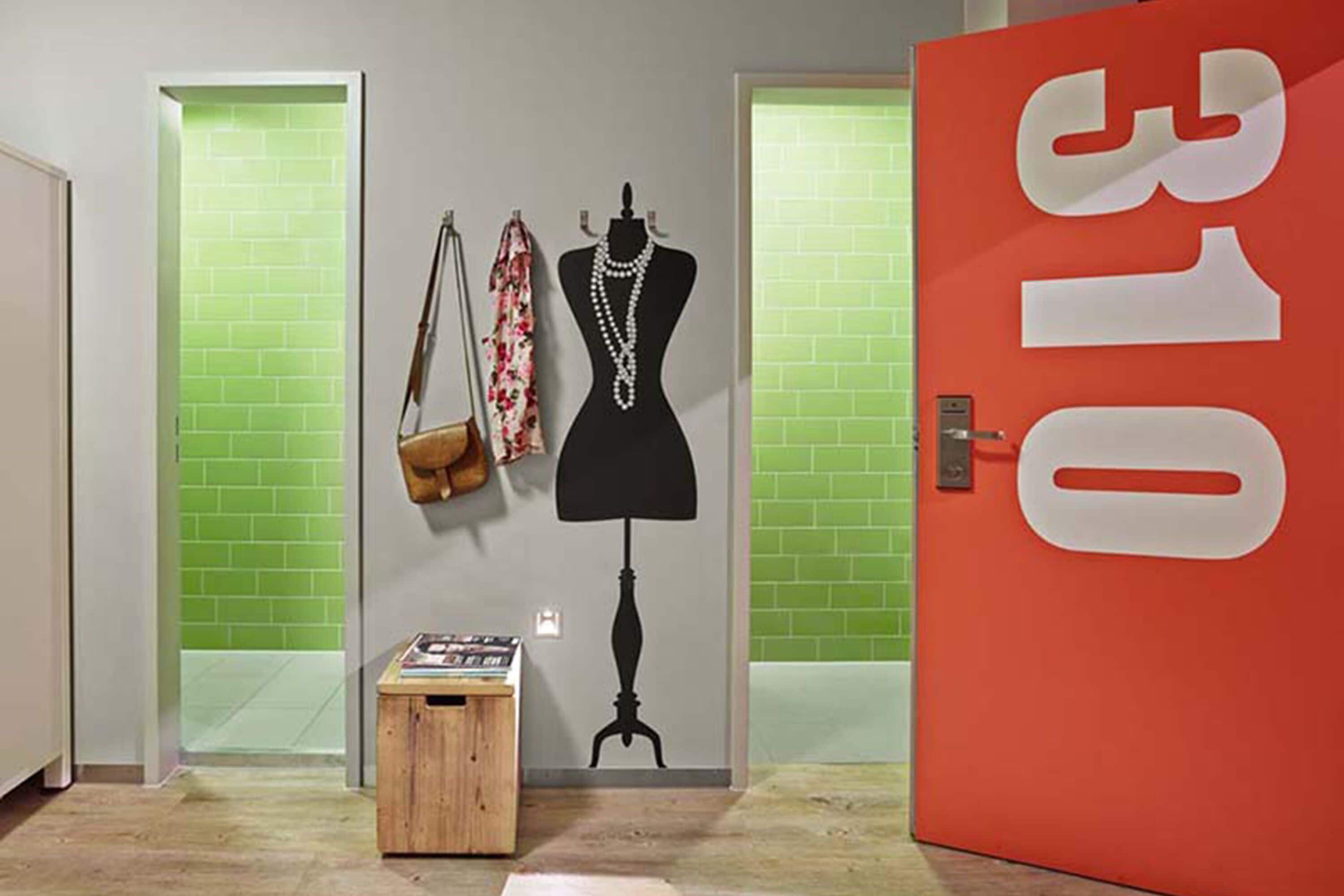 Una habitación con un maniquí vestido con un vestido negro, una bufanda estampada y un bolso de mano en una pared. Hay un taburete de madera pequeño con objetos encima. Se ve una puerta roja con el número 310.