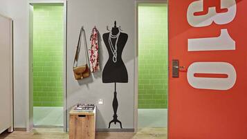 Una habitación con un maniquí vestido con un vestido negro, una bufanda estampada y un bolso de mano en una pared. Hay un taburete de madera pequeño con objetos encima. Se ve una puerta roja con el número 310.