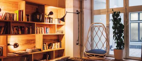 A cozy living room with a leather sofa, wooden bookshelves, a wicker chair, and a potted plant by a large window.