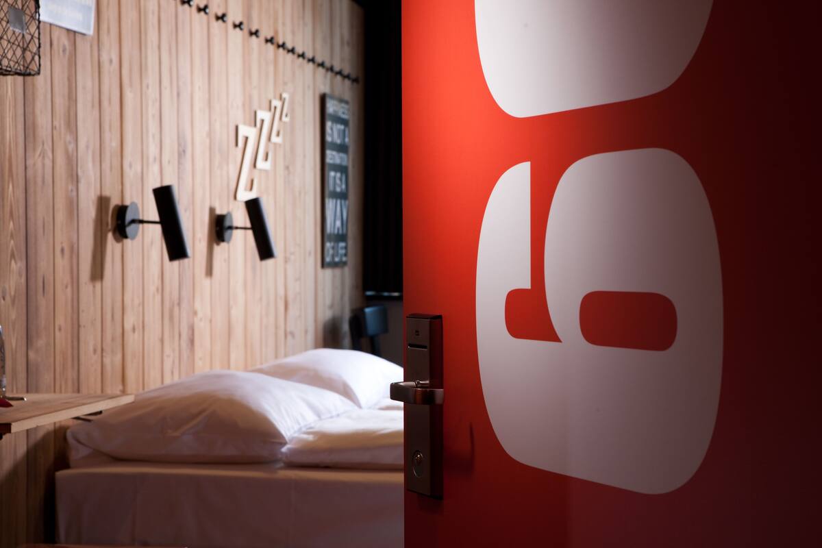 A hotel room with a wooden wall, a bed with white bedding, and a red accent wall with a large white logo.