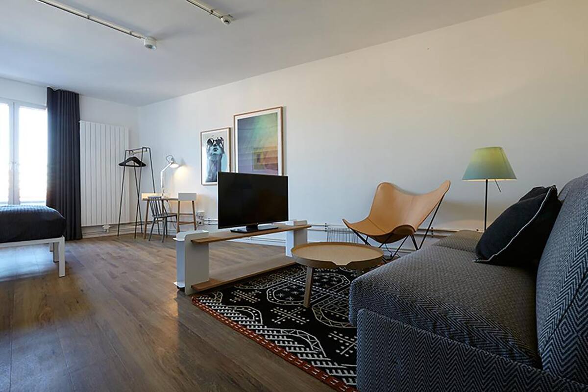 A modern hotel room with a bed, a TV, a sofa, a small table, and a chair. There is a window with curtains, a lamp, and a framed picture on the wall.