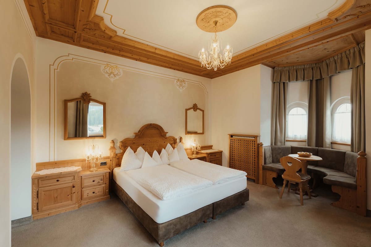 A bedroom with a large bed, wooden headboard, bedside tables, a chandelier, a mirror, a window with curtains, and a seating area with a chair and table.