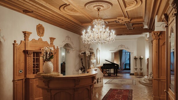 A grand lobby with a wooden reception desk, a chandelier, a grand piano, and a red patterned rug. - Hotel Colbricon  (Primiero San Martino di Castrozza)