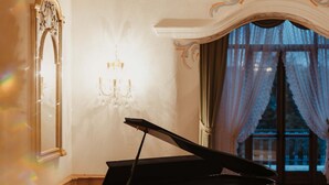 A grand piano in a well-lit room with a chandelier, a large mirror, and a view through a window with curtains. - Hotel Colbricon  (Primiero San Martino di Castrozza)
