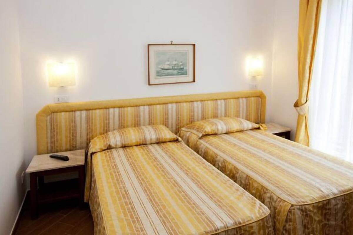 Une chambre d’hôtel avec deux lits simples, une table de chevet, un tableau encadré au mur et une fenêtre avec des rideaux.