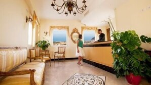 Ein Empfangsbereich mit Rezeptionist, Theke, einer Couch, Sesseln, einer Kristallleuchtre und einem großen Fenster mit Blick.