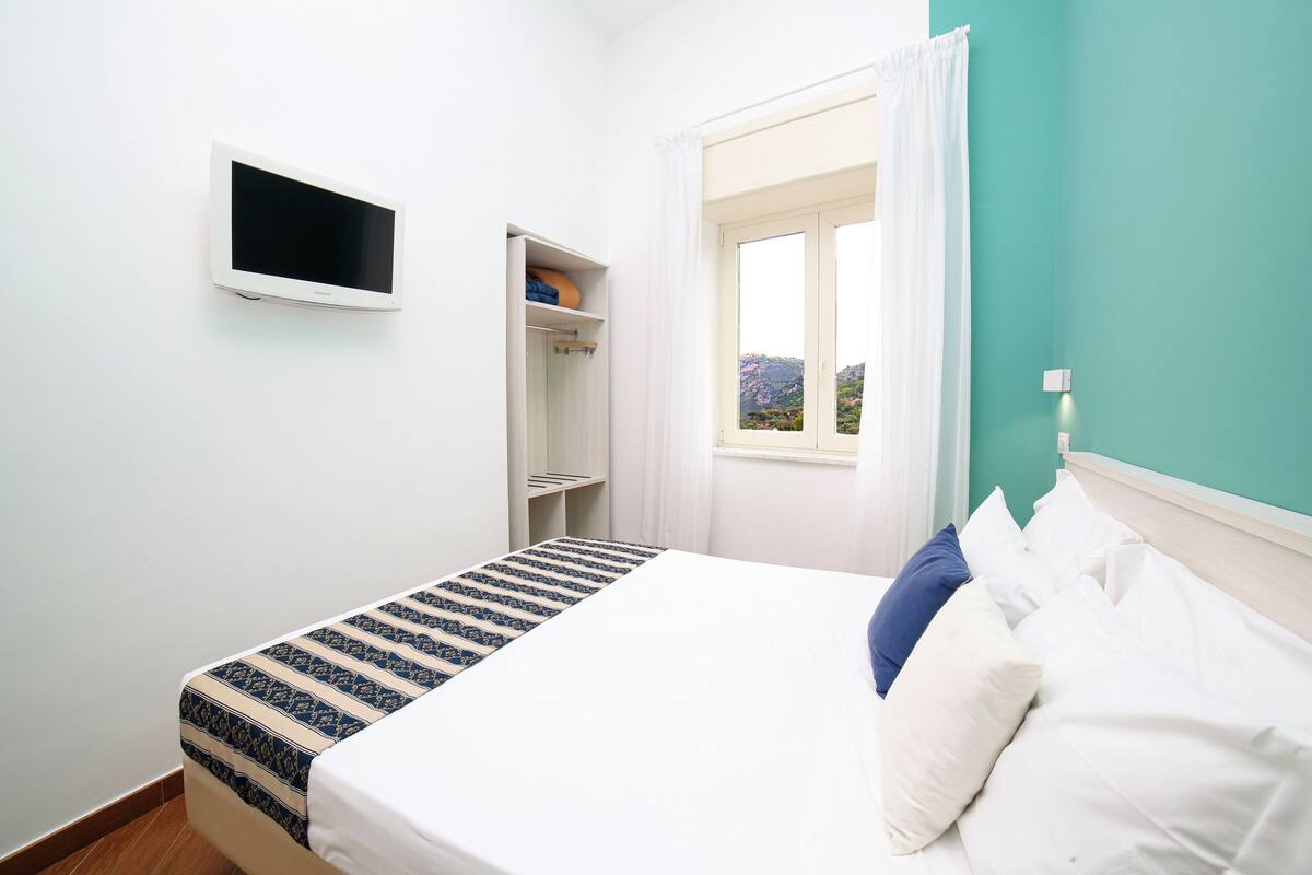 Une petite chambre à coucher, bien rangée, avec un lit, un téléviseur fixé au mur, une fenêtre avec des rideaux et une étagère sur laquelle se trouvent une serviette et un petit objet.