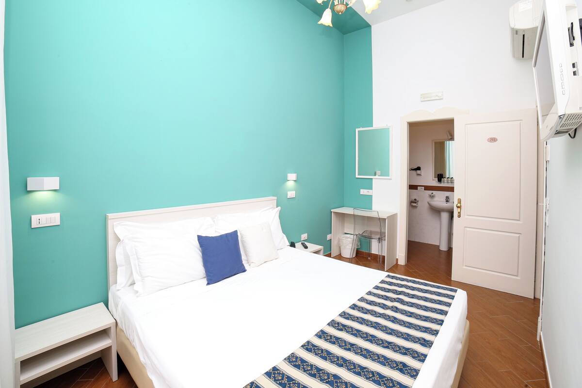 Une chambre à coucher comprenant un lit, une table de chevet, une commode, une salle de bain avec un lavabo et un miroir, et une porte.