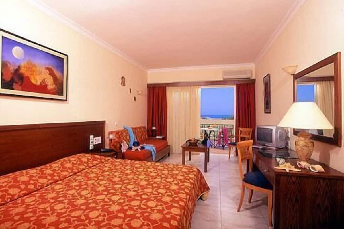 A hotel room with a large bed, a desk with a lamp, a television, and a view of the ocean.