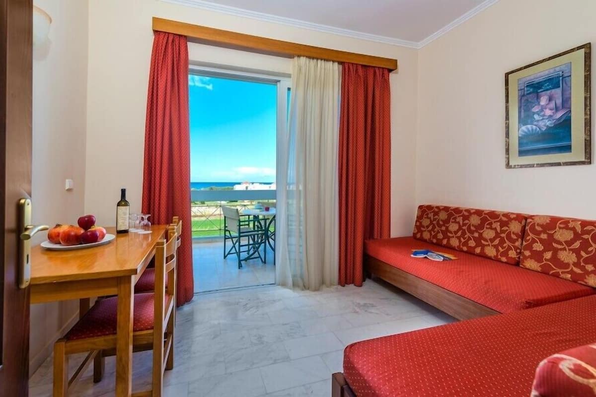 A room with a wooden table set with fruit and a bottle, a red sofa, and a view of the sea through a glass door.