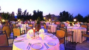 An outdoor dining area with round tables set for multiple guests, surrounded by chairs and tables covered with white tablecloths. - Selini Suites (Platanias)