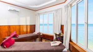 A room with two beds, a wooden wall, an air conditioning unit, a window with curtains, and a view of the sea.