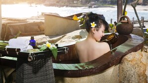 A person in a wooden tub with a towel and a flower, near a beach with boats and a rocky shore.