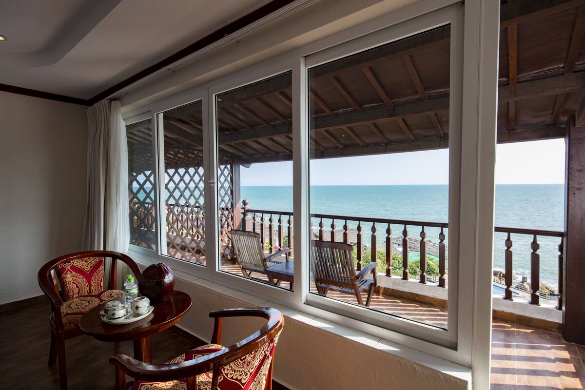 A balcony with a view of the sea, wooden chairs, and a table with a vase and teapots.