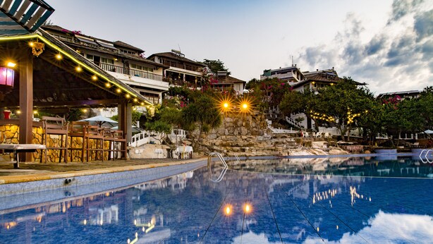 A swimming pool with a stone border, a covered seating area with tables and chairs, and a building with balconies in the background.