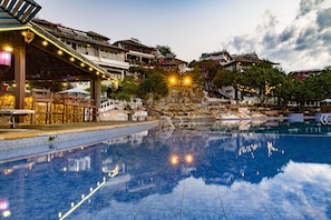 Une piscine bordée de pierres, un espace de détente couvert avec des tables et des chaises, et un bâtiment avec des balcons en arrière-plan.
