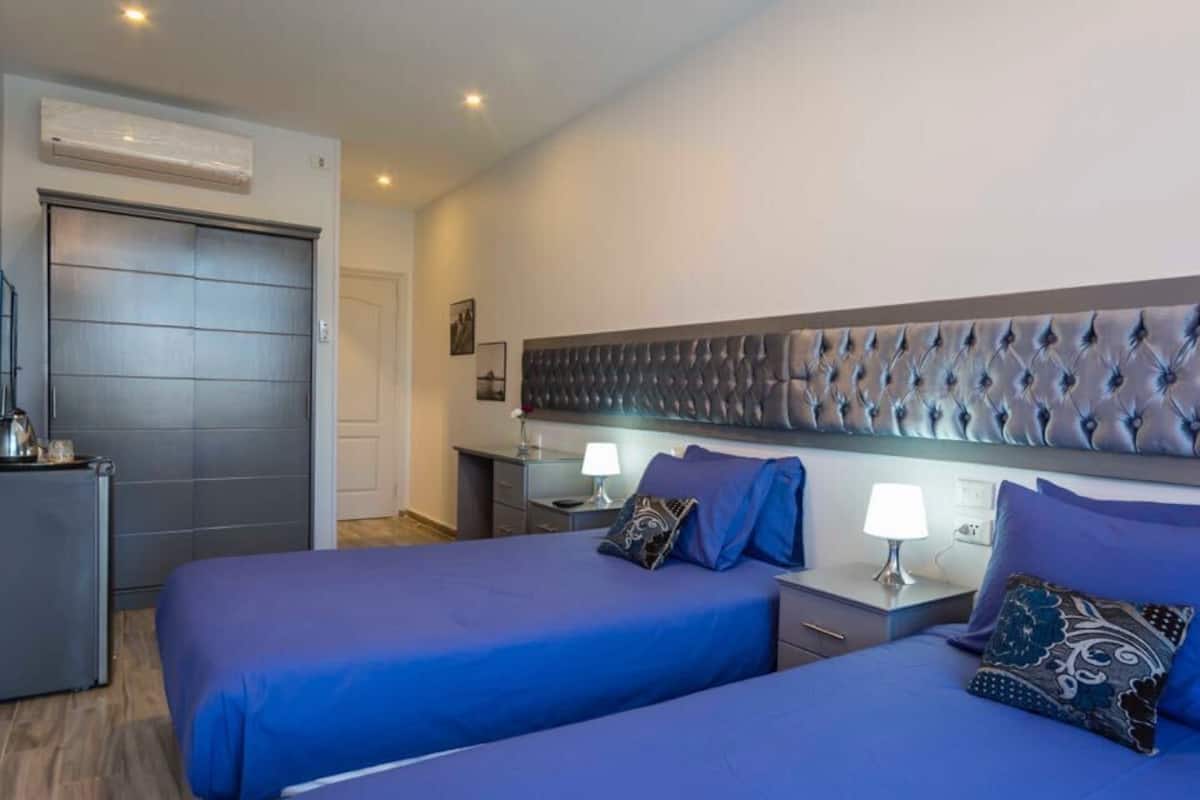 A hotel room with two beds, a large padded headboard, bedside tables with lamps, and a door with a handle.