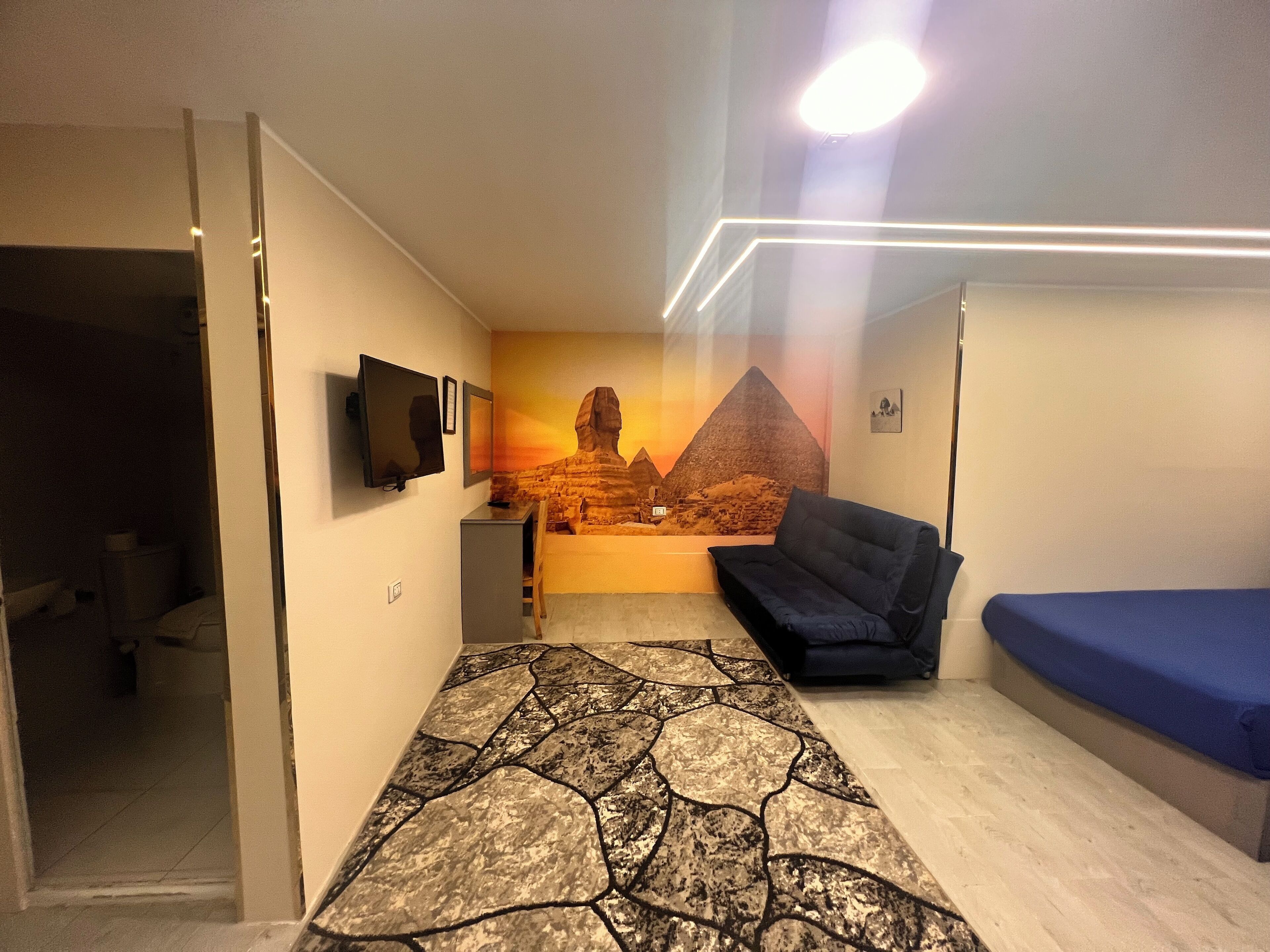 A room with a patterned carpet, a blue sofa, a bed with a blue mattress, a wall-mounted TV, and a mural of pyramids.