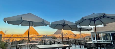 Un espace extérieur aménagé avec des tables, des chaises, des parasols et une vue sur les pyramides.