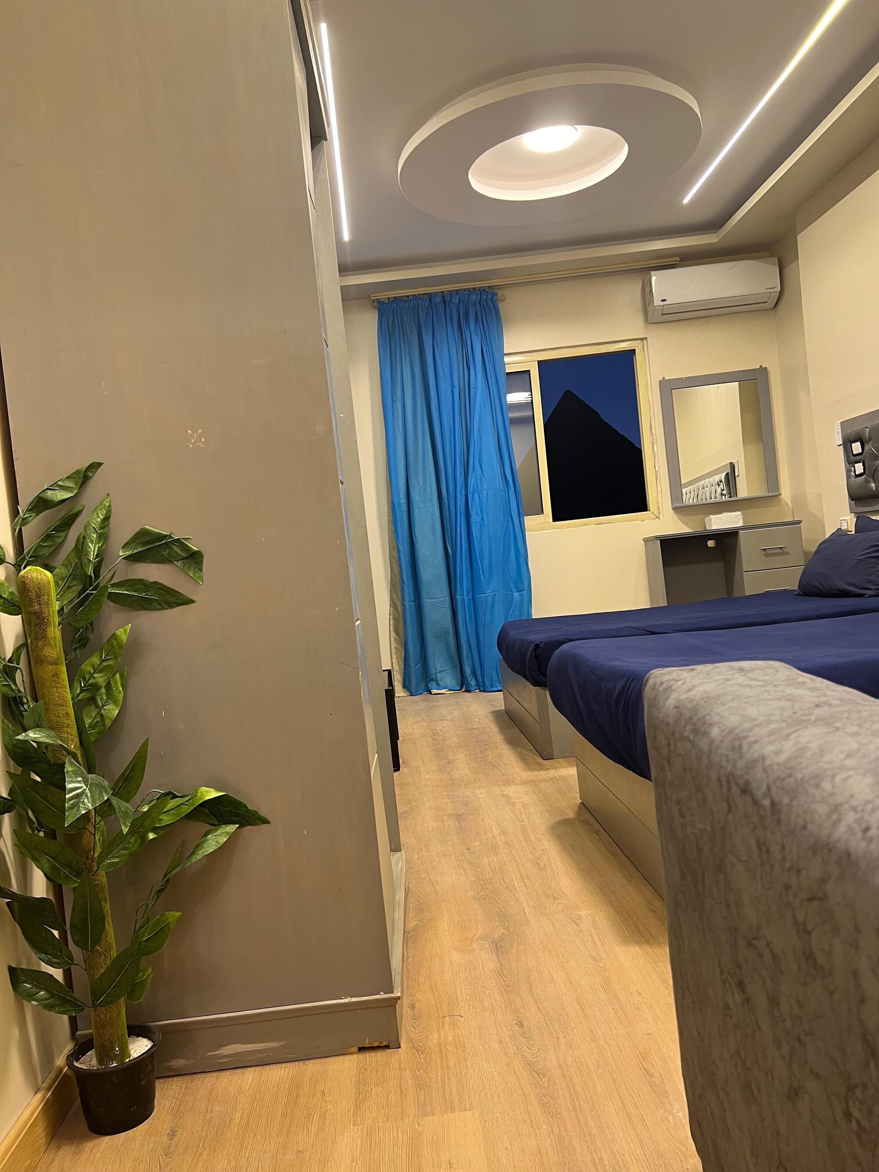 Habitación de hotel con cama, mesita de noche, espejo, planta en maceta y ventana con cortinas.