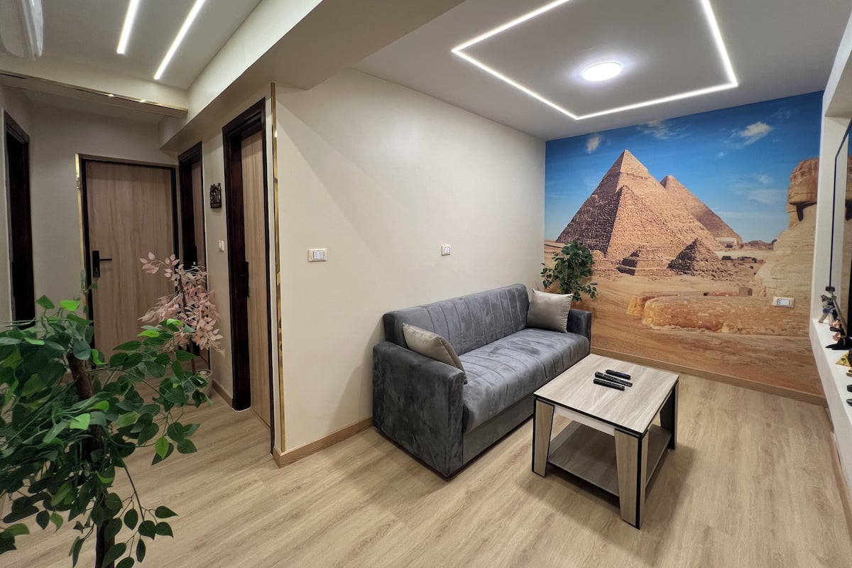 A modern living room with a grey sofa, a small wooden coffee table, and a wall mural of a pyramid.